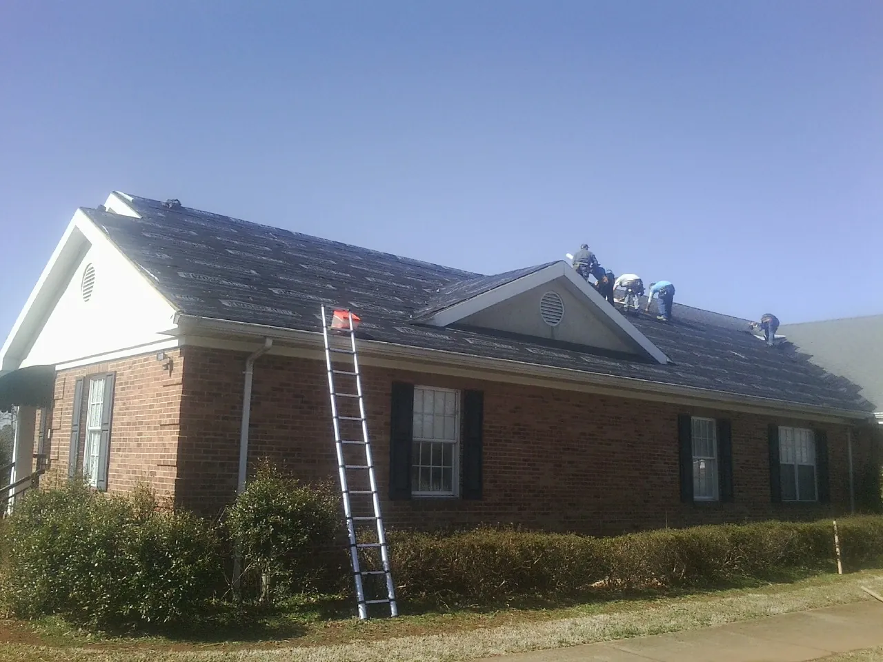Crew tearing shingles off a brick ranch roof with a ladder set — Emergency Roof Repair work by our crew in Roscoe